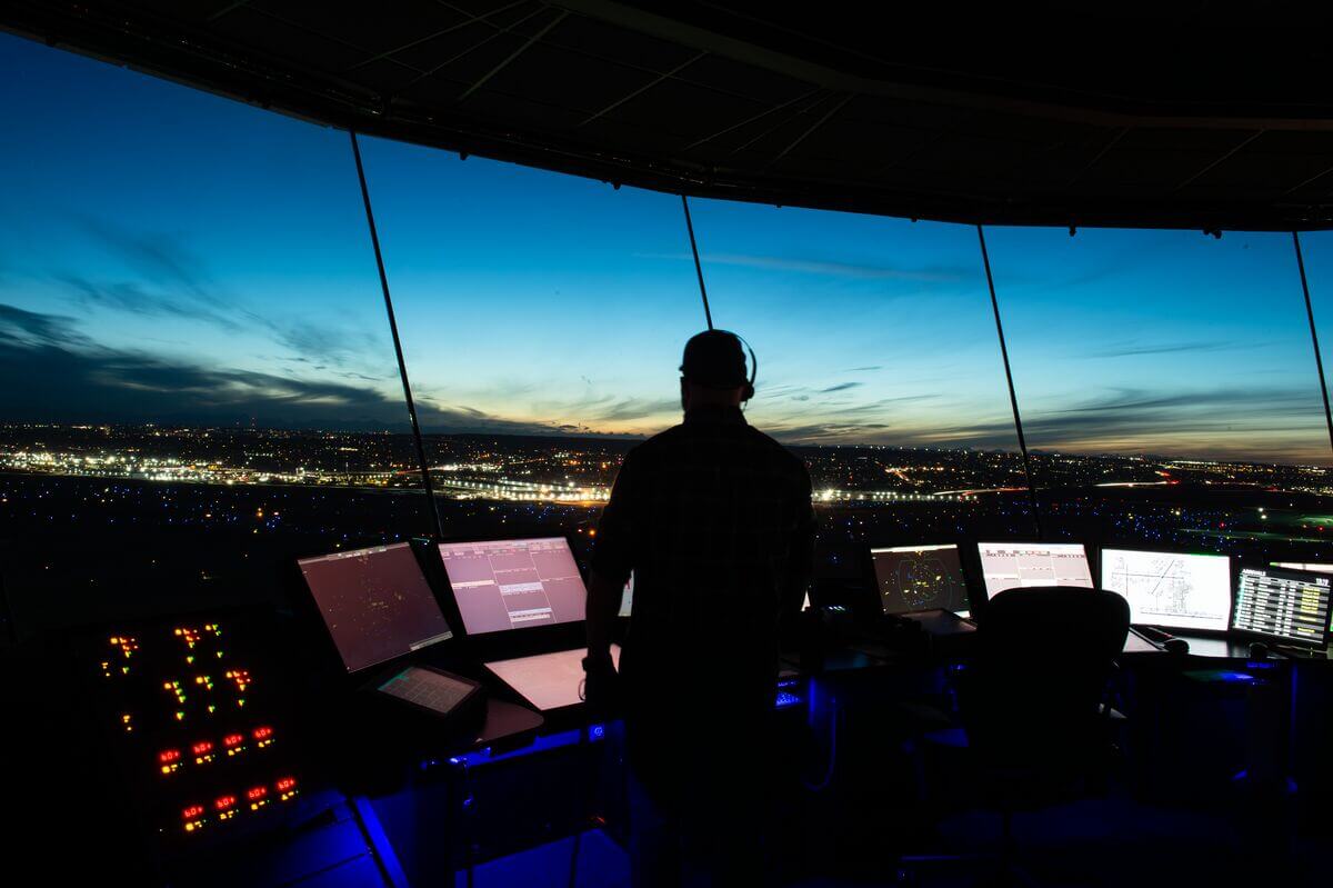 An air traffic controller stands in a control tower at dusk, overlooking a brightly lit airport.